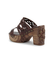 The Bed Stu Kreep is a 70's-inspired brown platform sandal featuring a natural wooden heel with floral cutout design on its straps, adorned by decorative metal studs along the edges.