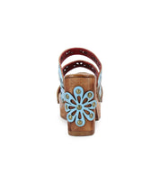 Bed Stu's Kreep sandal features a wooden heel adorned with a light blue floral cutout design, seen from the back. The red and blue detailed straps create a 70’s-inspired look.