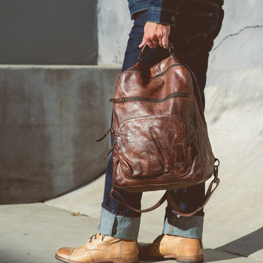 A man is holding a brown leather Bed Stu Lafe backpack.