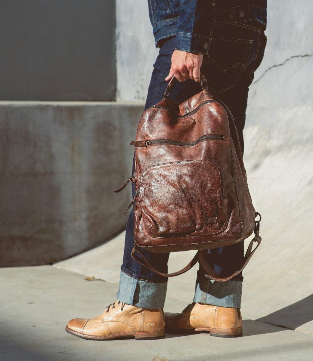 A man is holding a brown leather Bed Stu Lafe backpack.