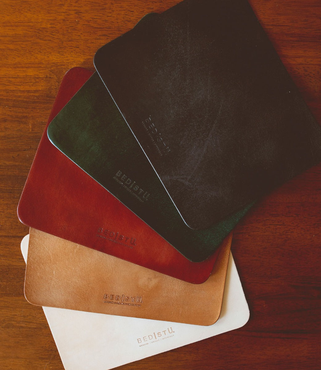 Five leather swatches in black, green, red, tan, and white are arranged in a fan pattern on a wooden surface within a curated workspace. The Launcher Mouse Pad has "Bed Stu" embossed on it.