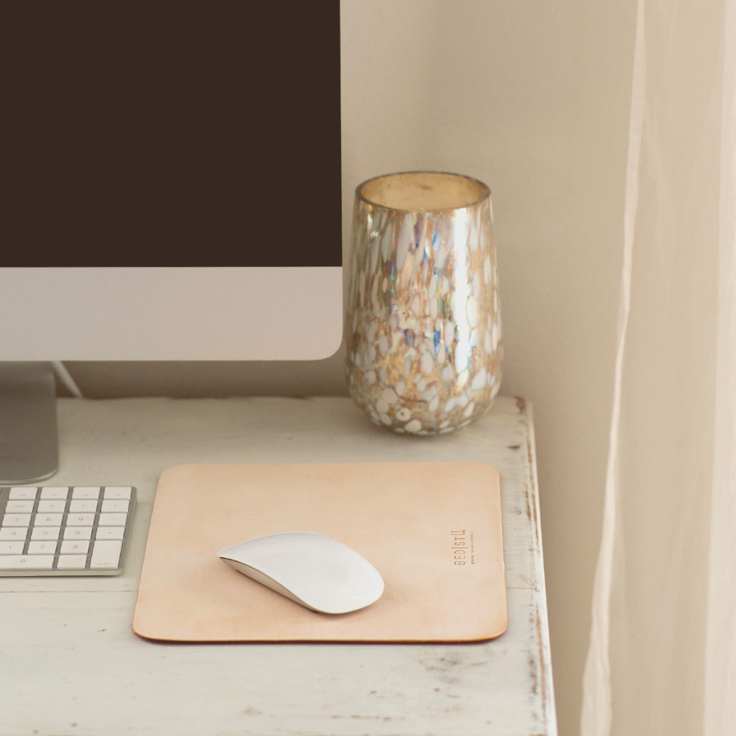 A computer monitor, keyboard, and mouse on a white wooden desk with a beige leather **Launcher Mouse Pad by Bed Stu** featuring a suede underside and a decorative cup create a curated workspace.
