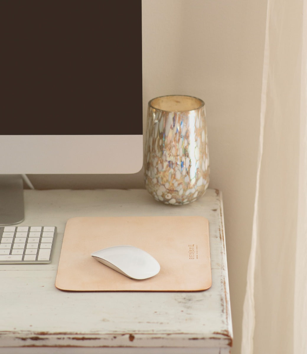 A computer monitor, keyboard, and mouse on a white wooden desk with a beige leather **Launcher Mouse Pad by Bed Stu** featuring a suede underside and a decorative cup create a curated workspace.