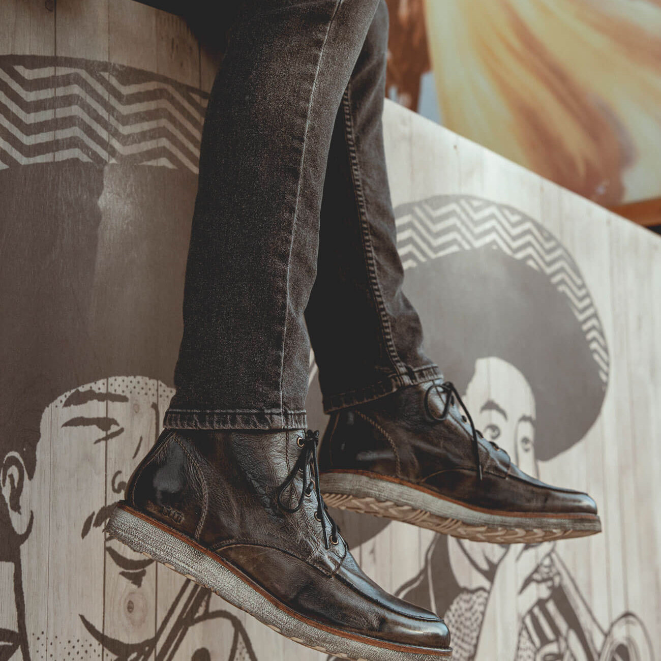 A person sitting on a ledge, wearing black jeans and Bed Stu men's leather boots, with a mural of a face in the background.