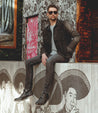 Man in sunglasses, denim jacket, and Bed Stu Lincoln work boots sitting on a wooden ledge in front of a wall with illustrated sombrero-wearing figures.