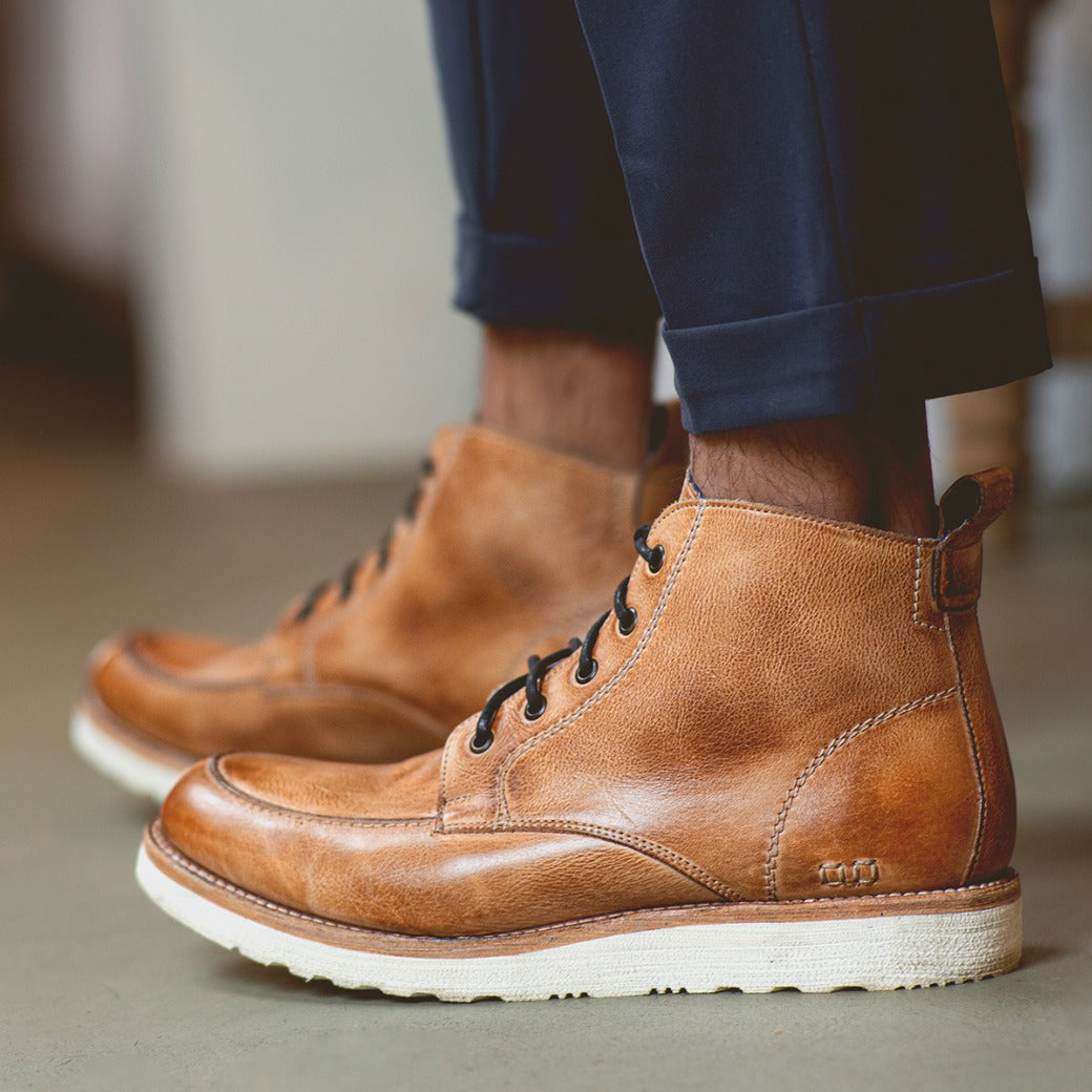 A man wearing brown Bed Stu men's leather boots and navy trousers, standing on a concrete floor.