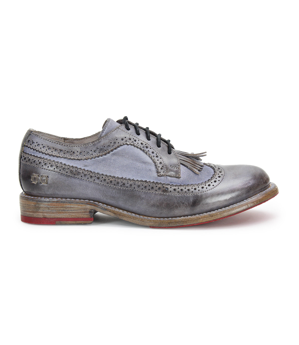 The Lita K by Bed Stu is a gray brogue shoe with a derby shape and black laces, featuring decorative perforations and a tassel on the tongue. It boasts a wooden sole with a red accent on the heel and a sophisticated wingtip toe.