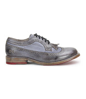 The Lita K by Bed Stu is a gray brogue shoe with a derby shape and black laces, featuring decorative perforations and a tassel on the tongue. It boasts a wooden sole with a red accent on the heel and a sophisticated wingtip toe.