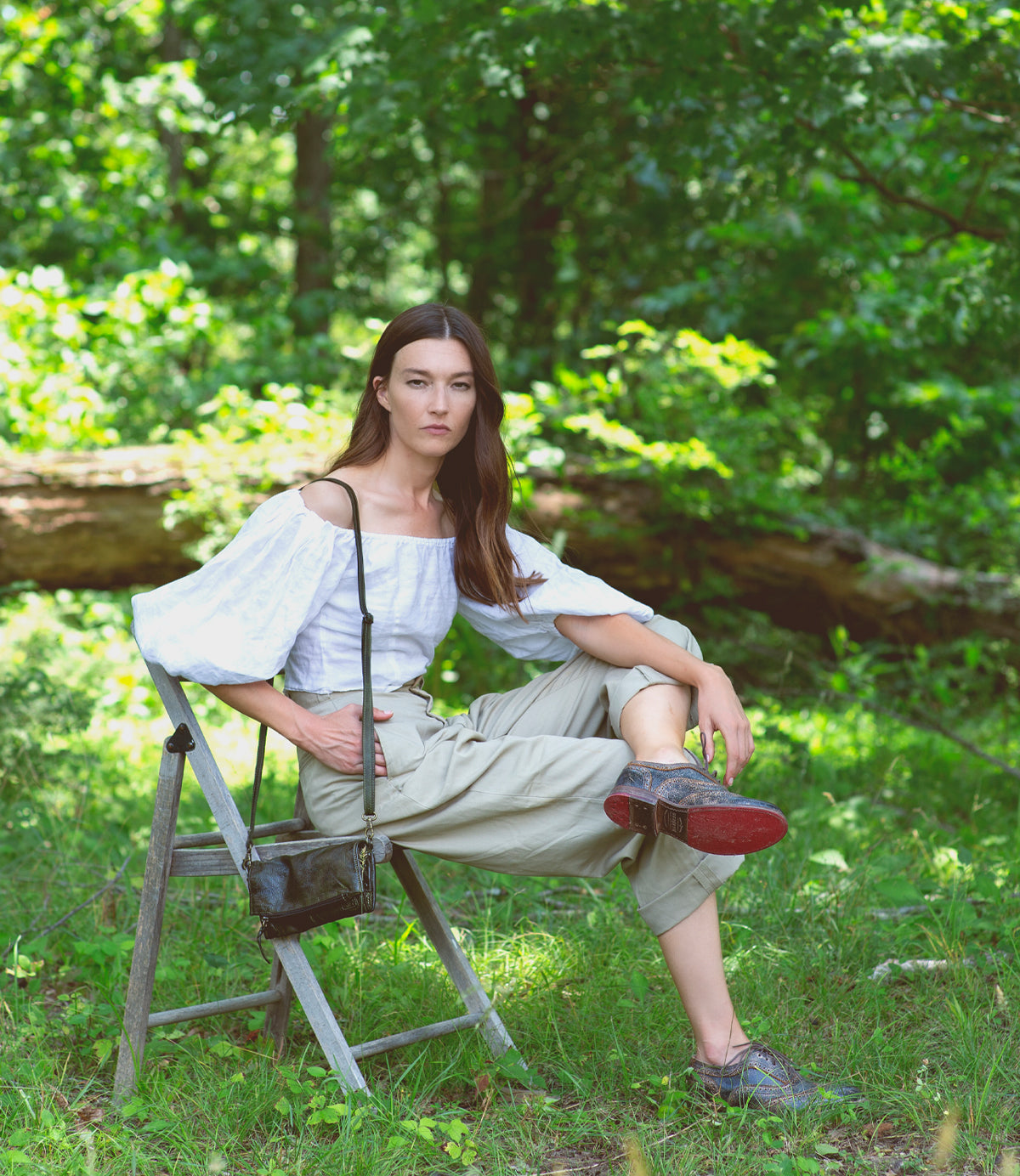 A woman sitting on a chair in the woods wearing a white top, distressed black Lita shoes and Amina bag.