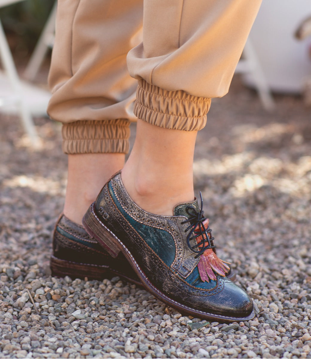 Person wearing tan jogger pants and Bed Stu Lita K vintage-style, multicolored leather oxford shoes with wingtip toe, standing on gravel.