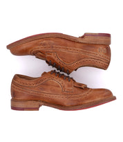 The Bed Stu Lita K is a pair of tan leather classic derby shoes with wingtip detailing, decorative perforations, stacked wooden soles, and red heel accents, displayed side by side on a white background.