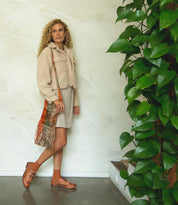 A woman with curly hair stands indoors near a leafy plant, wearing a beige jacket and skirt, orange socks, Bed Stu Lita K brown classic wingtip derby shoes, and carrying a fringed shoulder bag.