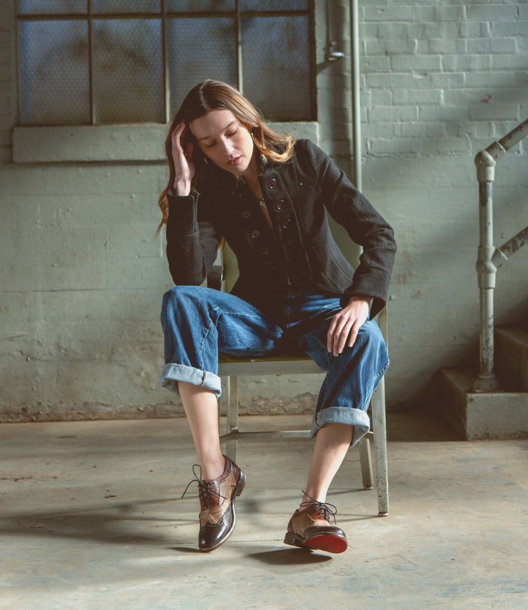 A woman sitting in a chair while wearing a black shirt, blue jeans, and brown and black Lita shoes.