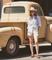 A woman in a cream hat, shorts, white shirt and tan Lita shoes standing next to an old truck.