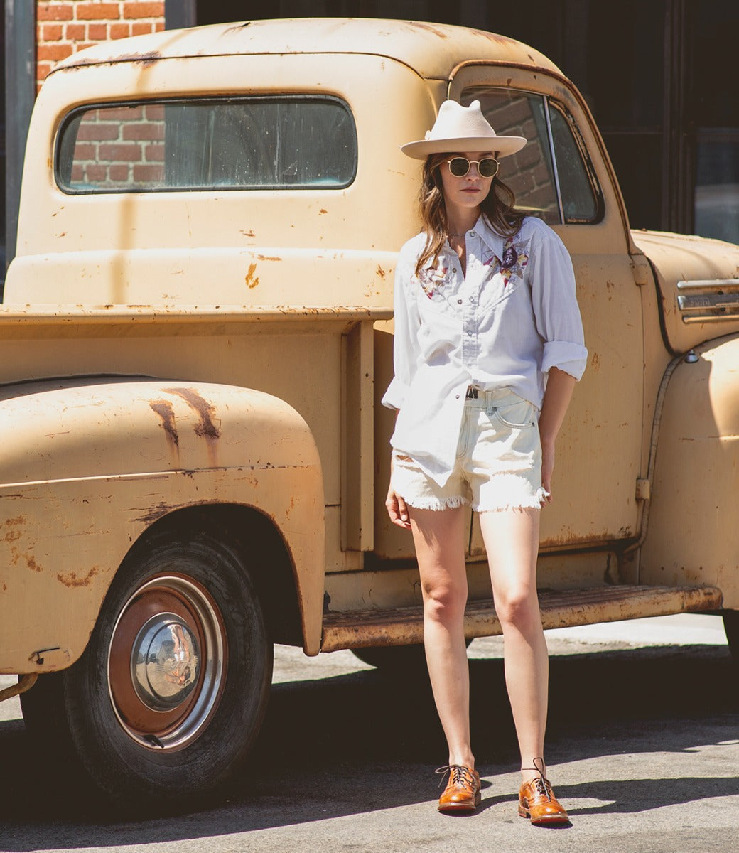 A woman in a cream hat, shorts, white shirt and tan Lita shoes standing next to an old truck.