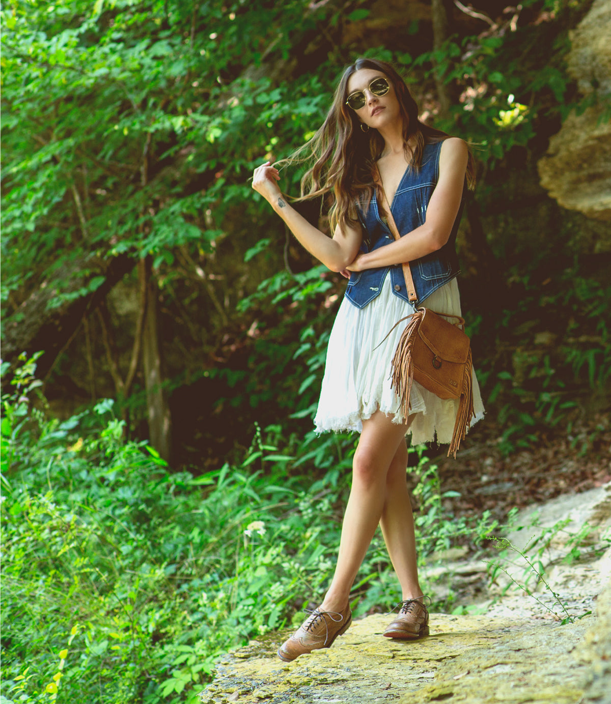 A woman in a denim vest, white dress, and tan Lita shoes and Hidden bag standing on a rock in the woods.