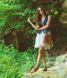 A woman in a denim vest, white dress, and tan Lita shoes and Hidden bag standing on a rock in the woods.
