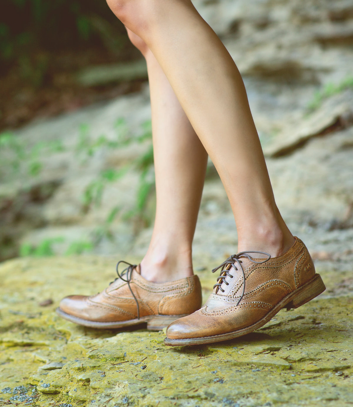 A woman's legs standing on a rock wearing tan Bed Stu Oxford shoes.