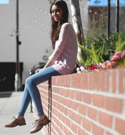 A woman sitting on a brick wall wearing brown Bed Stu Lita shoes and a pink sweater.