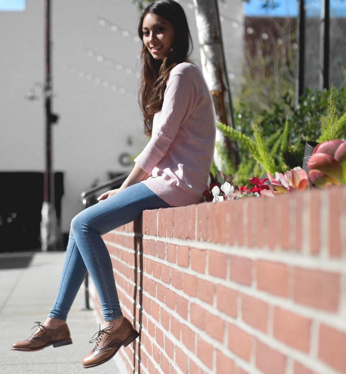 A woman sitting on a brick wall wearing brown Bed Stu Lita shoes and a pink sweater.