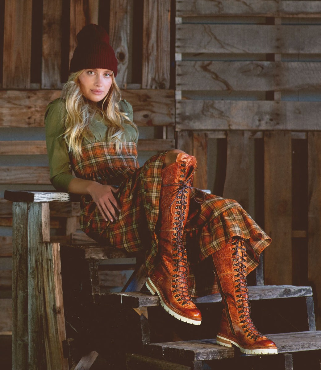 A woman wearing Lustrous boots and a hat sits on a wooden bench. Brand Name: Bed Stu.