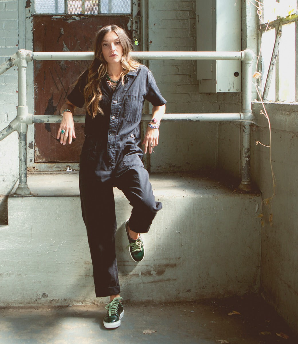 A woman in a black jumpsuit leaning against a railing, wearing Bed Stu's Lyne.