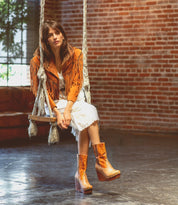 A woman in a fringed tan jacket and white dress sits on a swing wearing Bed|Stü Macarena color block leather wedge boots with a cushioned insole.