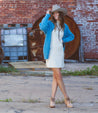 A woman in a cowboy hat, blue cardigan, white dress, and tan ankle boots standing in front of a brick building.