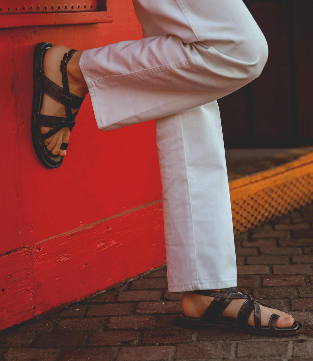 A person wearing Bed Stu Manati II pants and sandals leaning against a red wall.