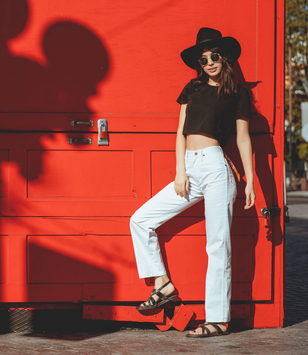 A woman leaning against a red wall wearing a Bed Stu hat and white jeans.