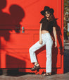 A woman leaning against a red wall wearing a Bed Stu hat and white jeans.