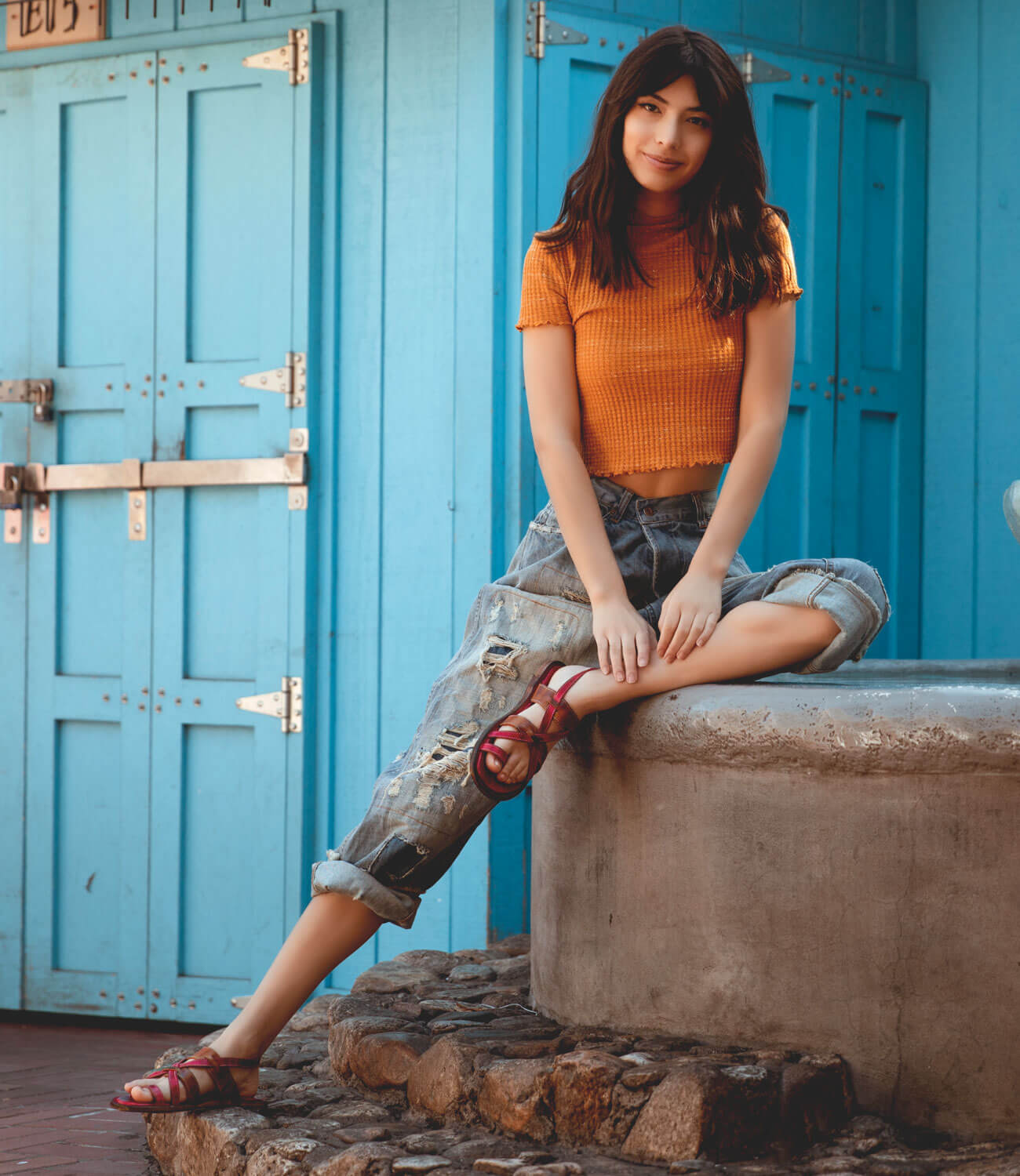 A woman in an orange top sitting on a blue wall wearing the Bed Stu Manati II.