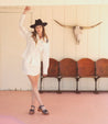 A woman in a white dress and cowboy hat posing in front of a pink wall with the Bed Stu Mantis.