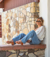 A woman sitting on a stone wall wearing Bed Stu jeans and a white shirt.