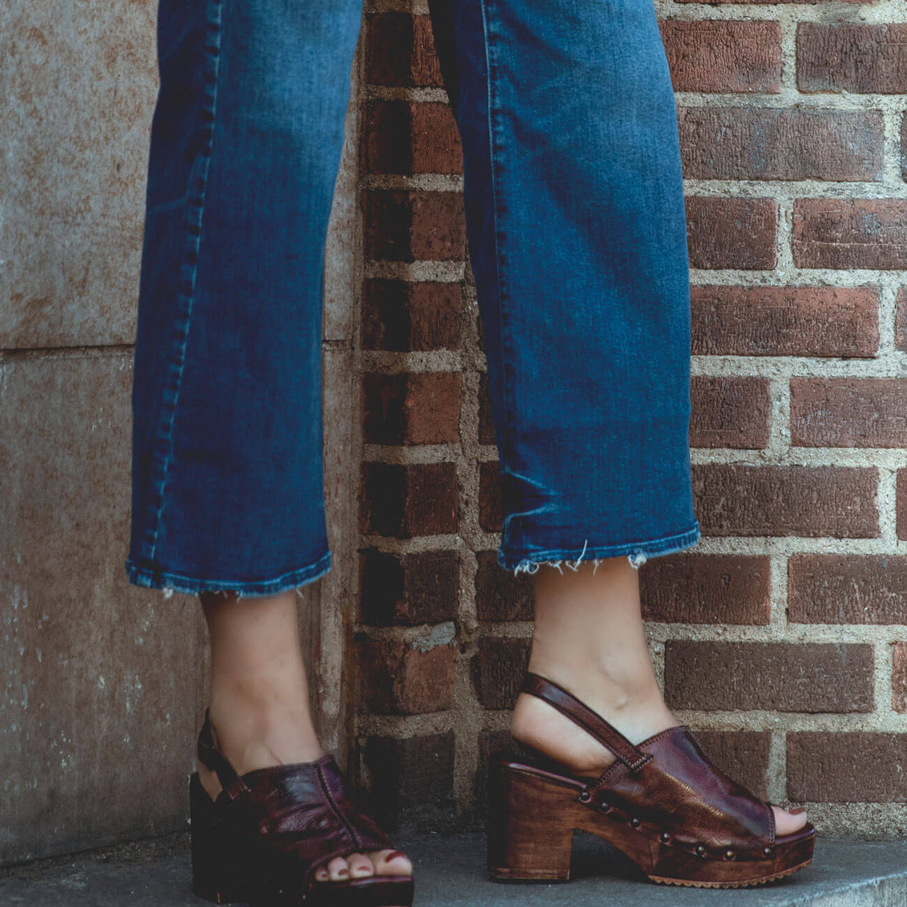 A woman wearing jeans and Marie sandals by Bed Stu on a brick wall.