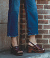 A woman wearing jeans and Marie sandals by Bed Stu on a brick wall.