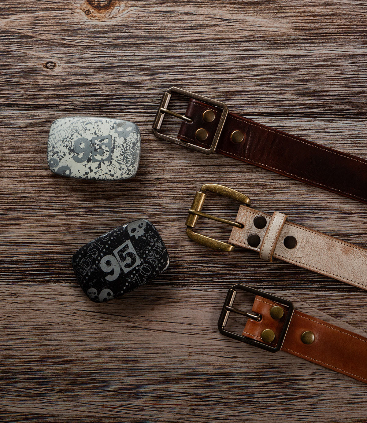 The Bed|Stü Meander Belt Buckle Bundle features two leather belts with brass buckles and two rectangular belt buckles, all displayed on a wooden surface.