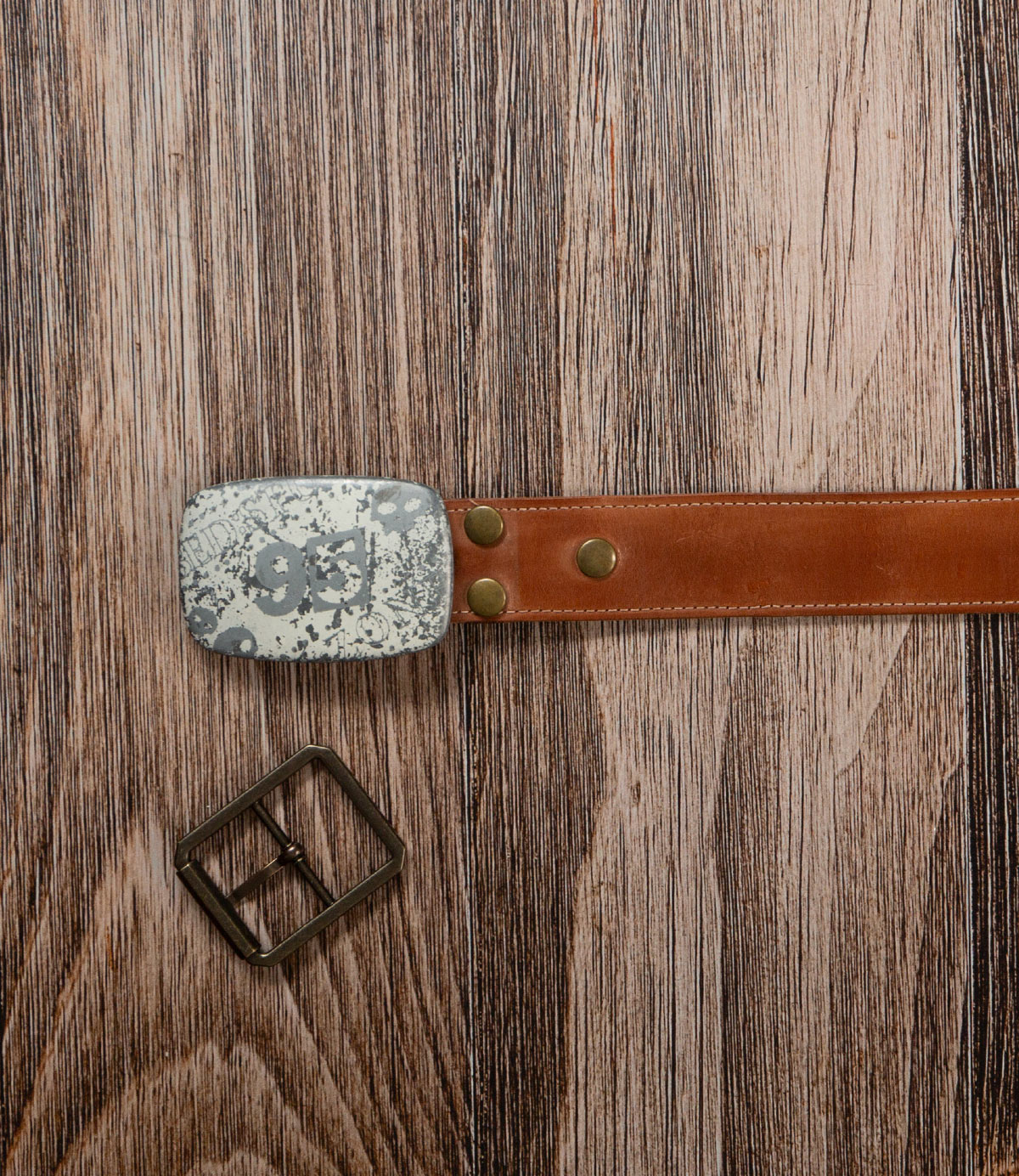 The Bed|Stü Meander Belt Buckle Bundle features a brown leather belt with a stylish gray patterned buckle, displayed on wood beside an extra metal belt buckle.