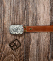 The Bed|Stü Meander Belt Buckle Bundle features a brown leather belt with a stylish gray patterned buckle, displayed on wood beside an extra metal belt buckle.