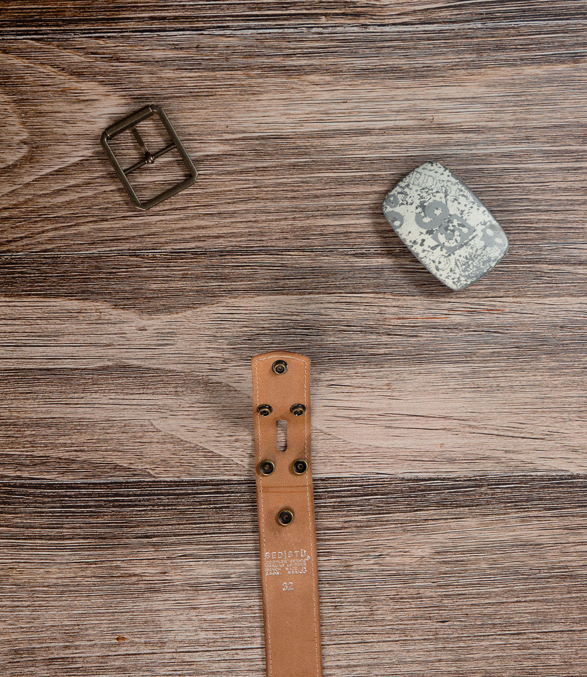 The Bed|Stü Meander Belt Buckle Bundle, featuring a disassembled brown leather belt, metal buckle, and silver belt plate, is displayed on a wooden surface.