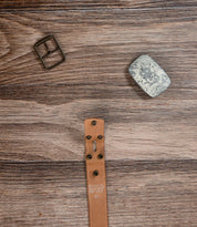 The Bed|Stü Meander Belt Buckle Bundle, featuring a disassembled brown leather belt, metal buckle, and silver belt plate, is displayed on a wooden surface.