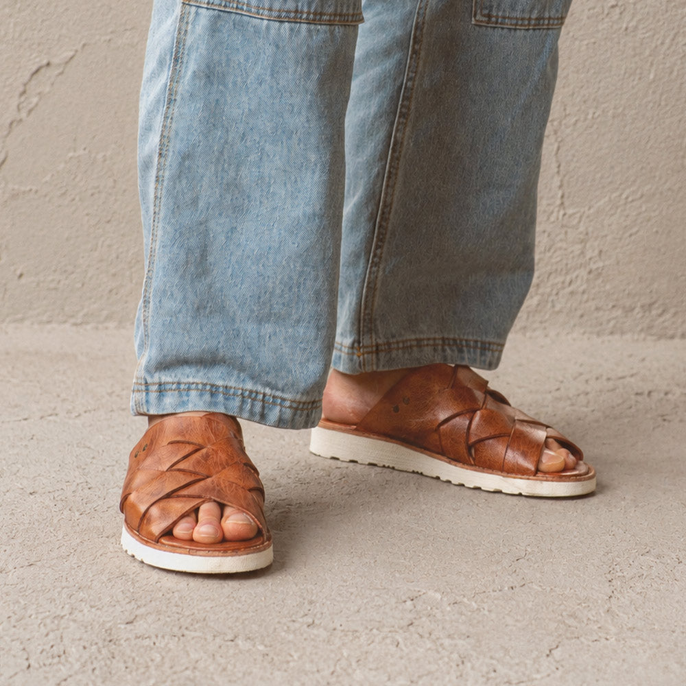 Brown woven leather slide sandals with open toes and white soles, worn with wide-leg light-wash jeans.