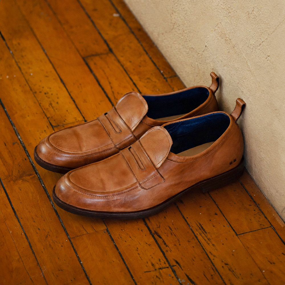 Pair of tan leather penny loafers with moc-toe stitching, dark soles, blue interiors, and heel pull tabs, resting on a wooden floor.