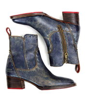 The "Merryli" ankle boots by Bed Stu, featuring worn blue leather with red tabs, side zippers, cushioned insoles, and stacked wooden heels, are showcased against a white background.
