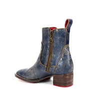 The Bed Stu Merryli ankle boot, shown in a side view, features distressed blue leather with a low wooden heel, side zipper, and pull tab. It also incorporates an eco-friendly cushioned insole for ultimate comfort.