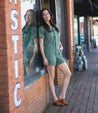 A woman leaning against a brick wall in a green Bed Stu overall romper named Mickie.