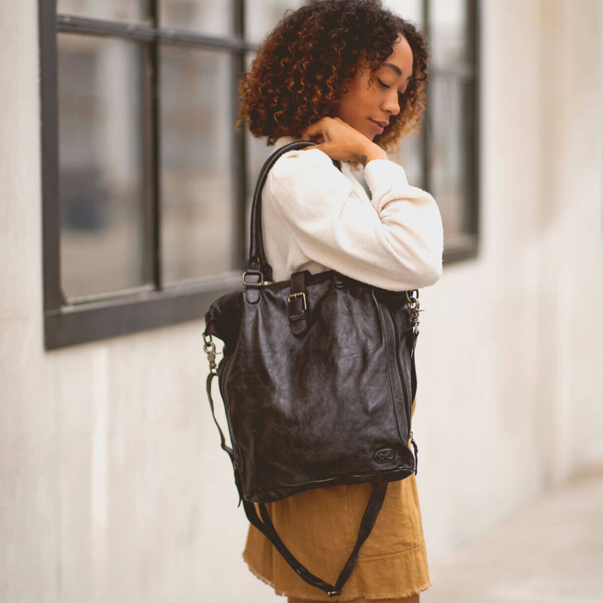 A woman stands outdoors wearing a white long-sleeve top and brown skirt, holding the Bed Stu "Mildred" large black leather tote bag with a crossbody strap over her shoulder.