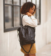 A woman stands outdoors wearing a white long-sleeve top and brown skirt, holding the Bed Stu "Mildred" large black leather tote bag with a crossbody strap over her shoulder.