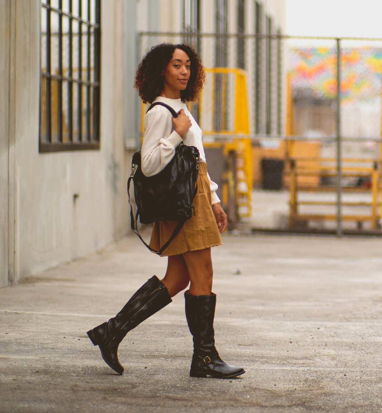 A person with curly hair walks outdoors, dressed stylishly and carrying the Bed Stu Mildred sleek leather tote bag. Industrial buildings and a fence are visible in the background.