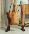 A woman holding a Mildred by Bed Stu brown leather tote bag.